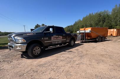 Woodland Rolloff black pickup truck towing an orange rolloff dumpster on a gravel lot